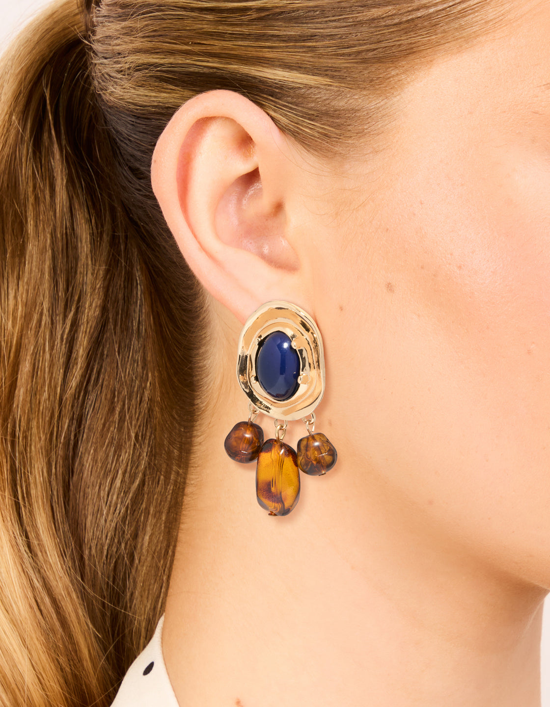 Showcasing statement charm and beaded medallion drop earrings in gold with midnight blue acrylic and saddle brown accents on ear view
