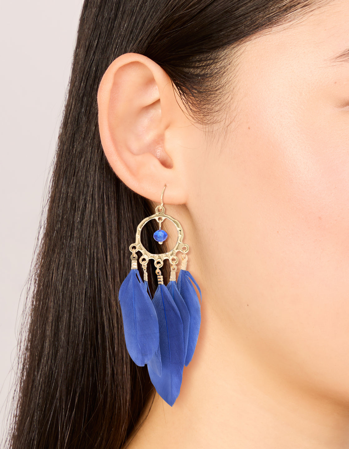Showcasing statement dreamcatcher earrings with beads, charms and dark blue feathers in a gold finish on ear view